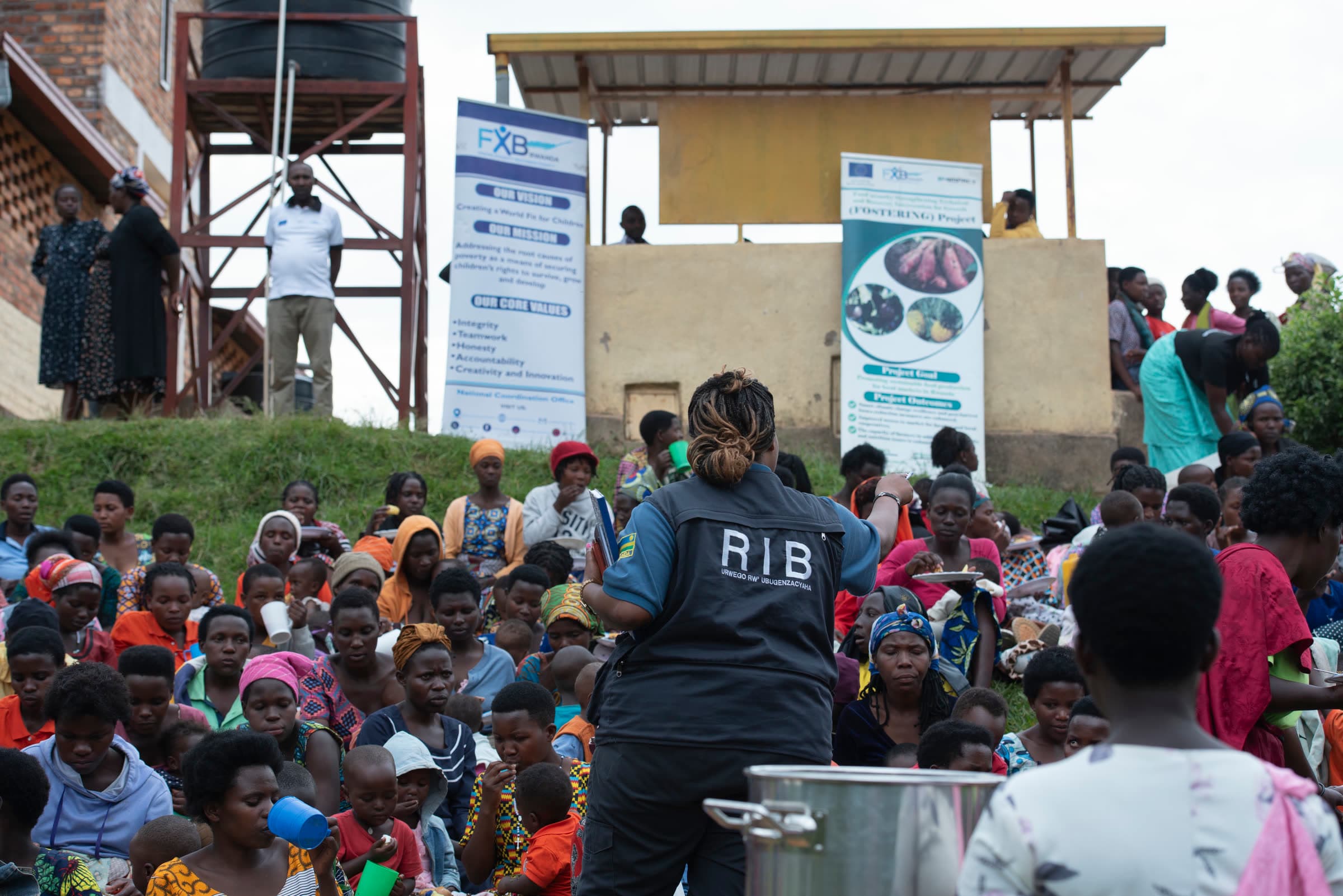 Women and children seated on the grass at a community gathering, in front of FXB Rwanda banners